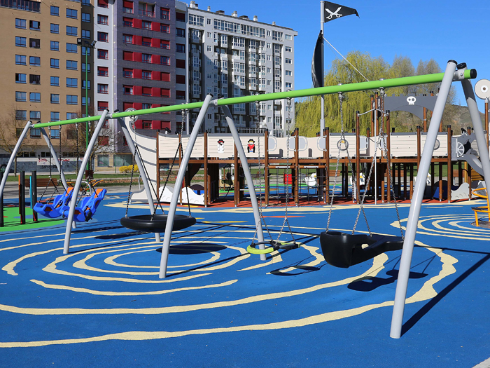 A three bay swingset with curved grey legs and a green crossbar situated in a playground next to a large residential block. In the background is a large play structure themed as a pirate ship. The play surface is blue with white markings to resemble water.
